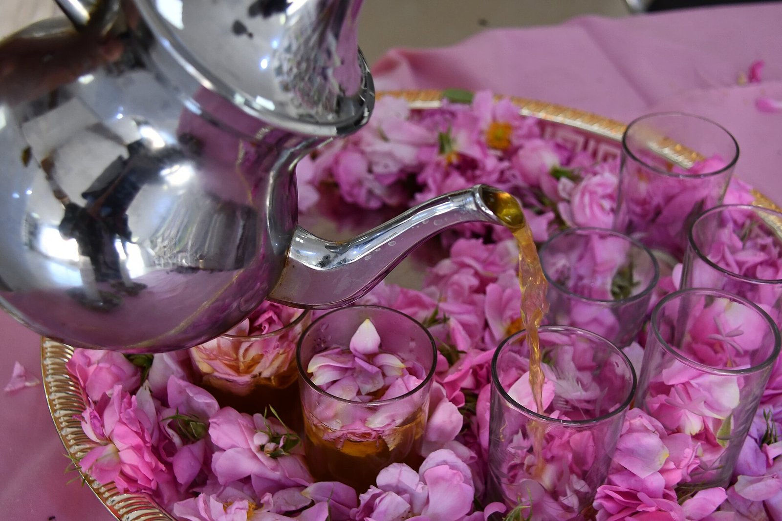 A silver teapot pouring mint tea into glasses among pink rose petals